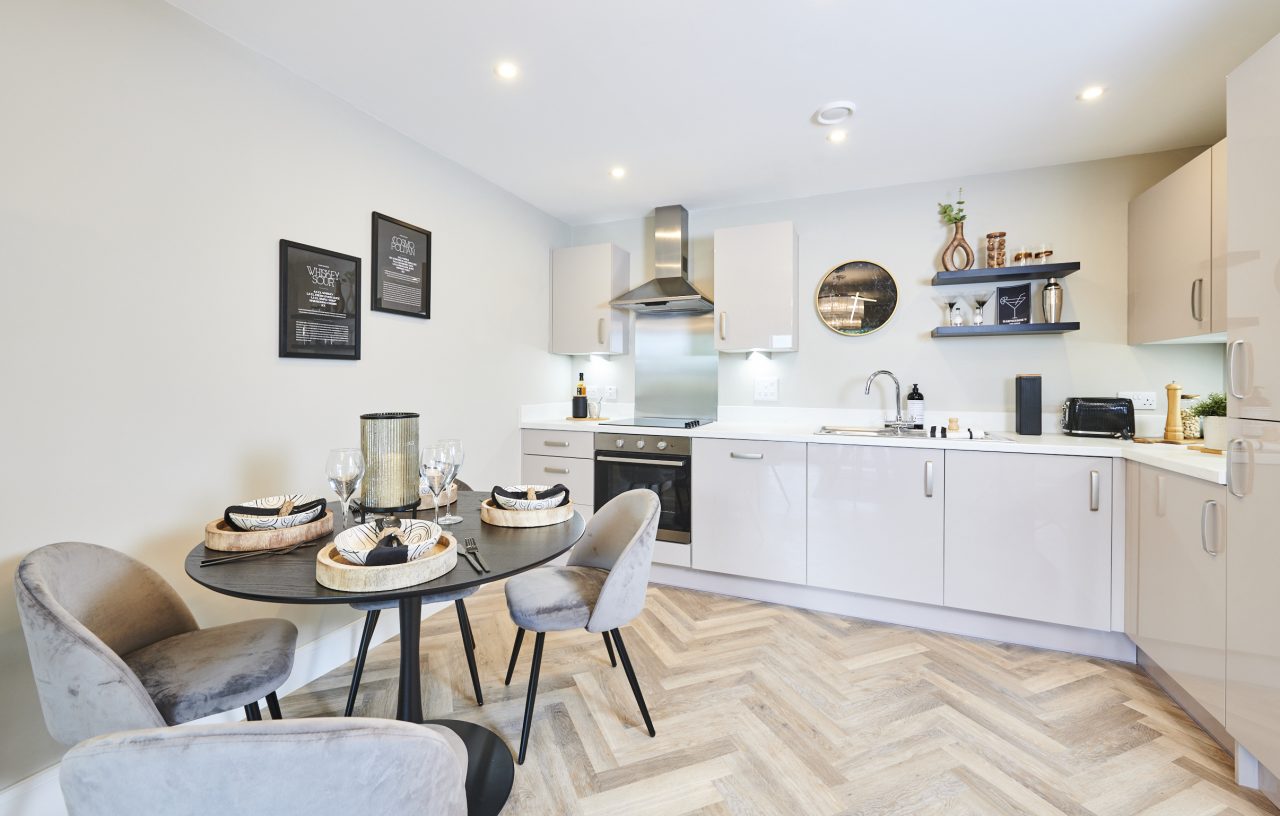 A kitchen dining room with herringbone flooring, almond cabinets with silver handles, and stainless steel hob and hood. Decorated with black and white modern prints, a black round table, and light beige coloured chairs with black legs.
