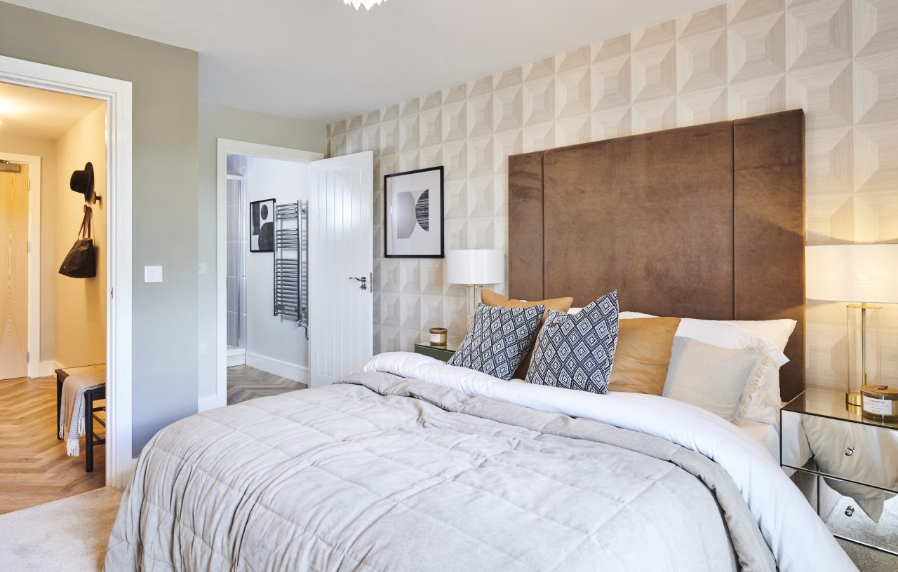 An interior shot of the new St Modwen Show Home in Austin view, this double bedroom has cream geoemetric textured wallpaper, a brown suede-look headboard and pillows three deep in grey, gold and stone.