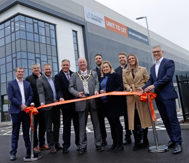 St. Modwen Logistics agents and Councillor Rod Laight with golden necklace on, standing in front of a newly completed warehouse, cutting an orange ribbon.